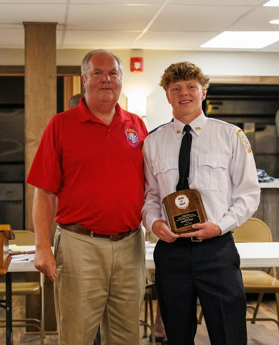 Two men stand indoors; one wearing a red polo shirt and khaki pants, the other in a white uniform shirt and tie, holding a plaque. They are posing and smiling for a photo at an award ceremony.